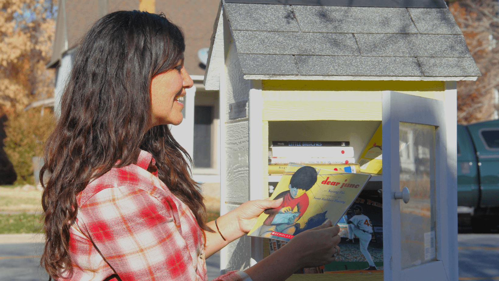 A woman taking a book out of a little library and smiling at the book.