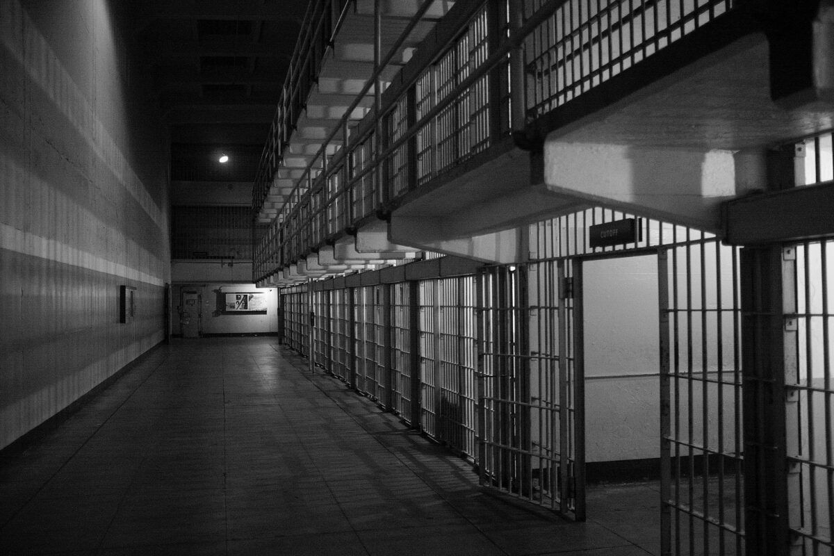 A black and white photo of a open and empty prison cells running down a dark hallway. 