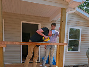 Two volunteers cutting a board together on a porch