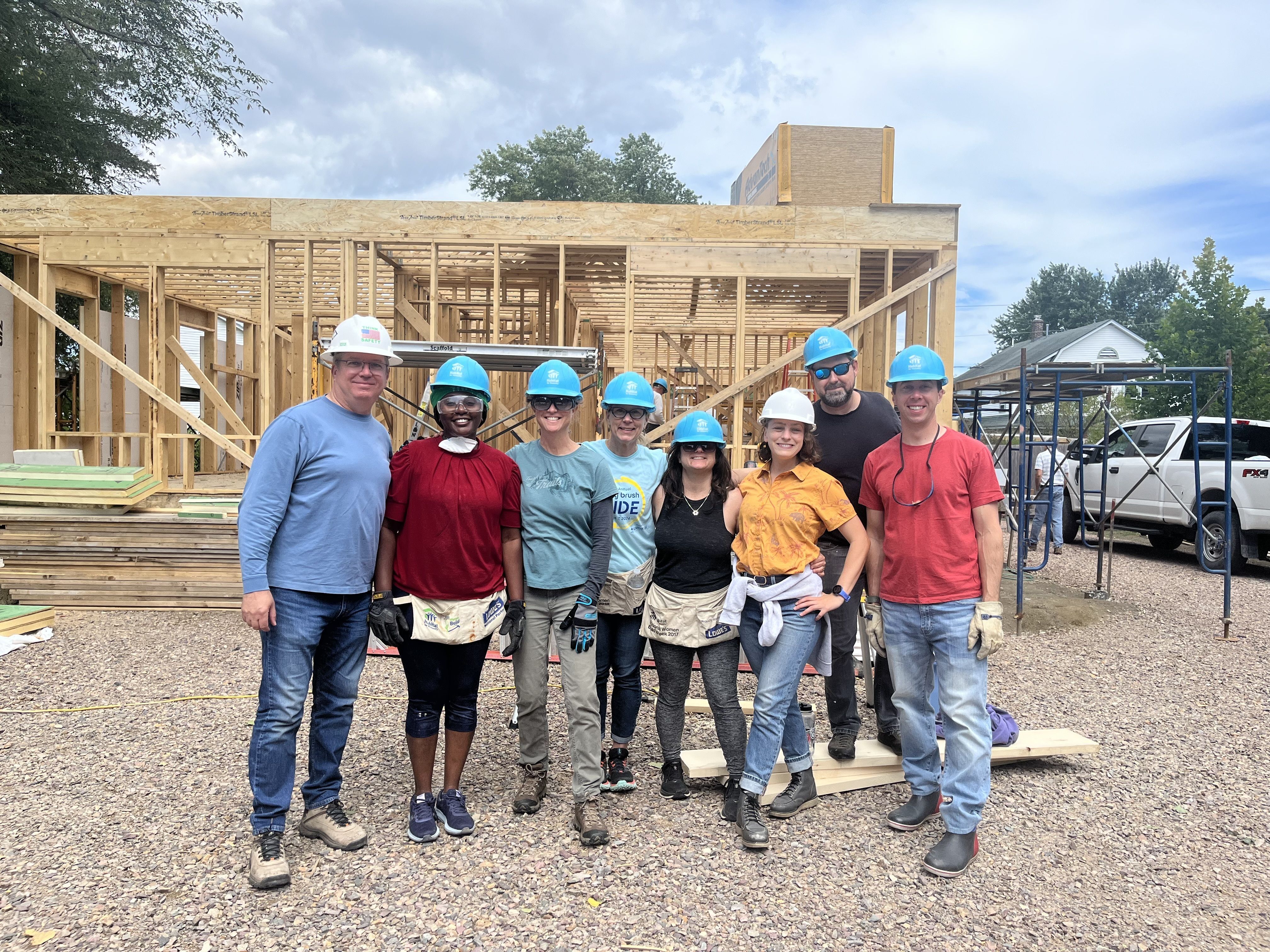 A group of volunteers wearing hard hats stands in front of a Habitat home that is currently being constructed.