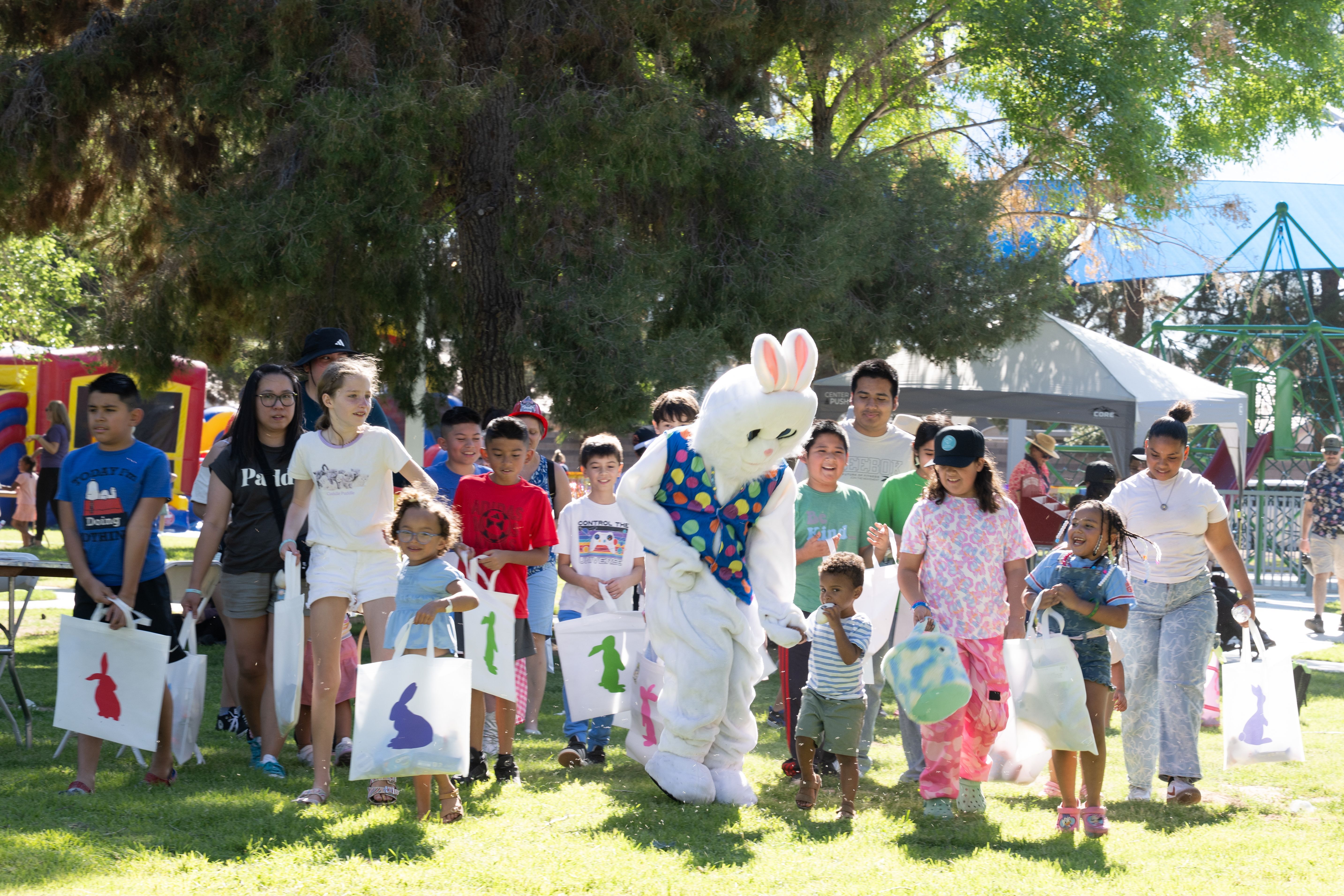 Children and families walk through a park alongside a costumed Easter bunny, carrying decorated bags and smiling during a festive outdoor egg hunt event.