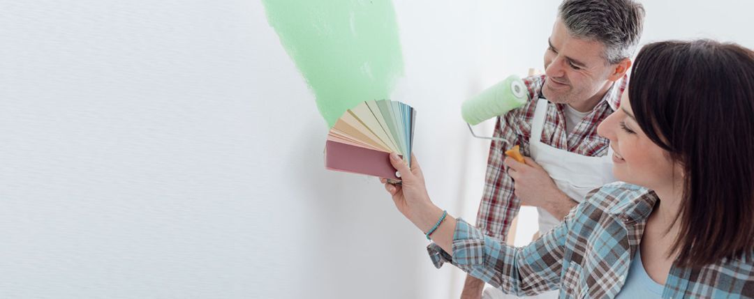 Couple choosing paint colors from a swatch while painting a wall with light green GreenSheen eco-friendly paint from Habitat ReStore Zephyrhills