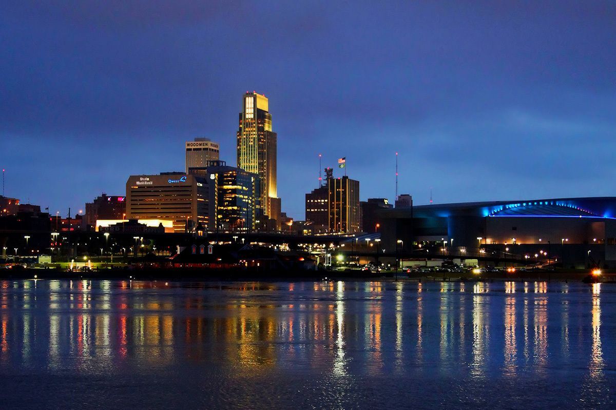 A nighttime image of the Omaha skyline from across the river in Iowa with many building lights reflecting across the water