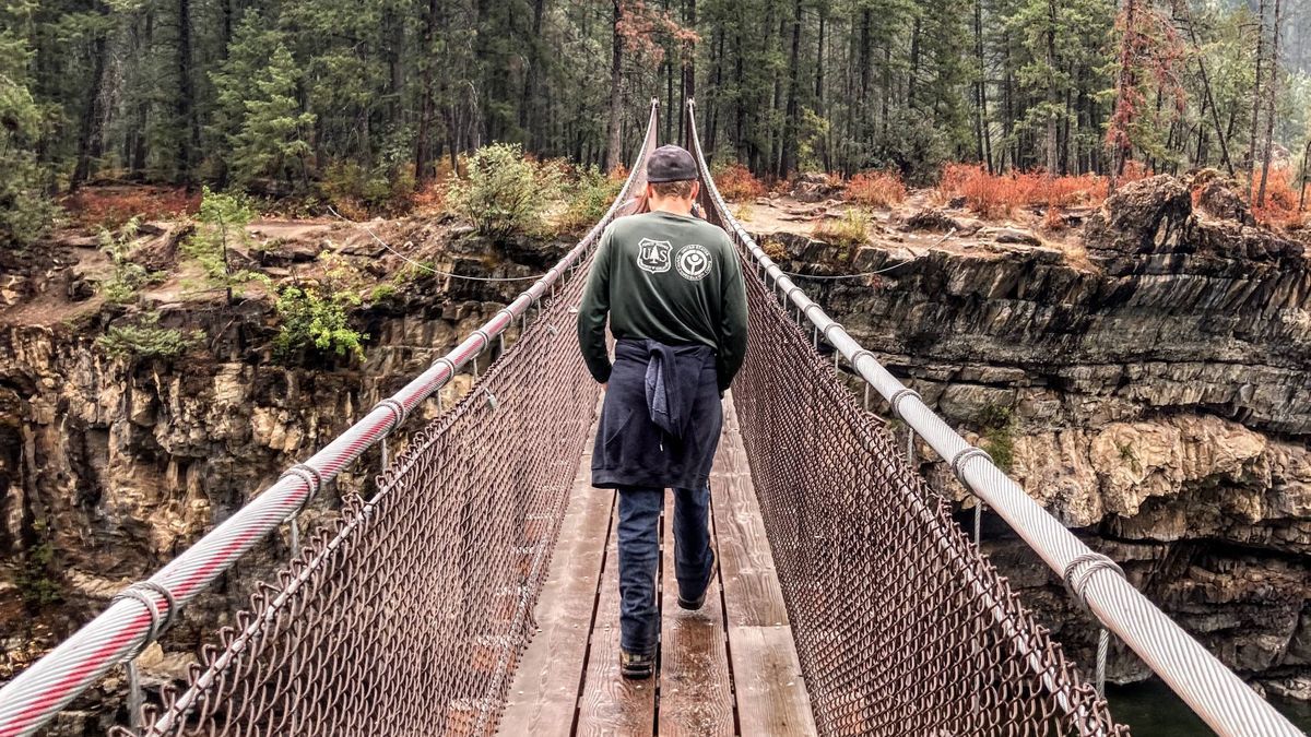 [Image Description: An MCC member is seen walking away from the camera with their back facing the viewer, walking a suspension bridge that connects between two deep forests.]