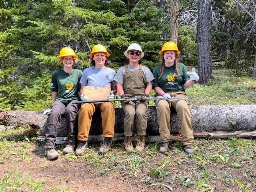 Three crew members sit on a log, holding tools and smiling