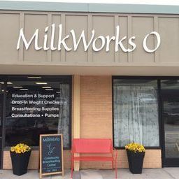The front of the MilkWorks in Omaha. There are yellow mums in black pots on either side of a red bench. The curtains are open and there is vinyl text on the window that says "Education & Support, Drop in Weight Station, Breasting Supplies, Consultations,