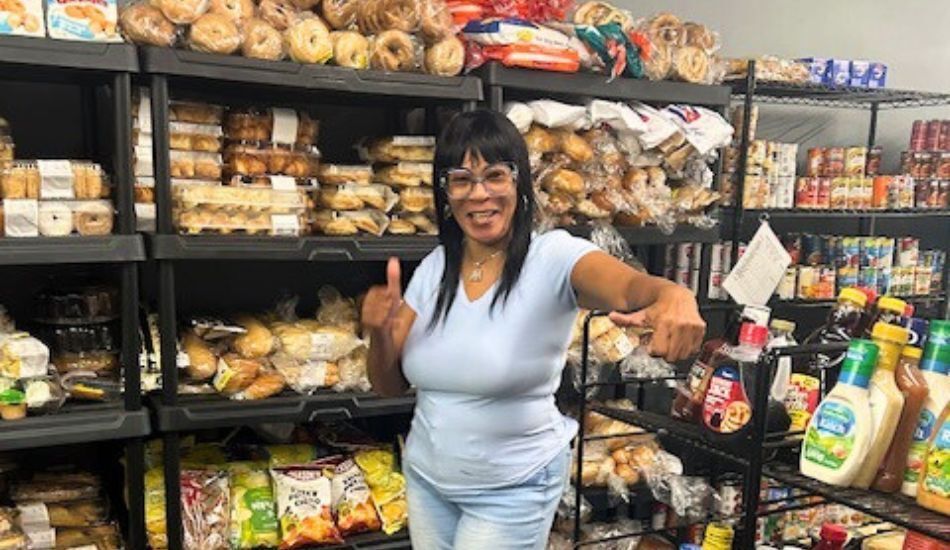 Photo of a smiling volunteer inside the food pantry. Shelves packed with pastries, canned goods, and shelf-stable food line the wall behind her.