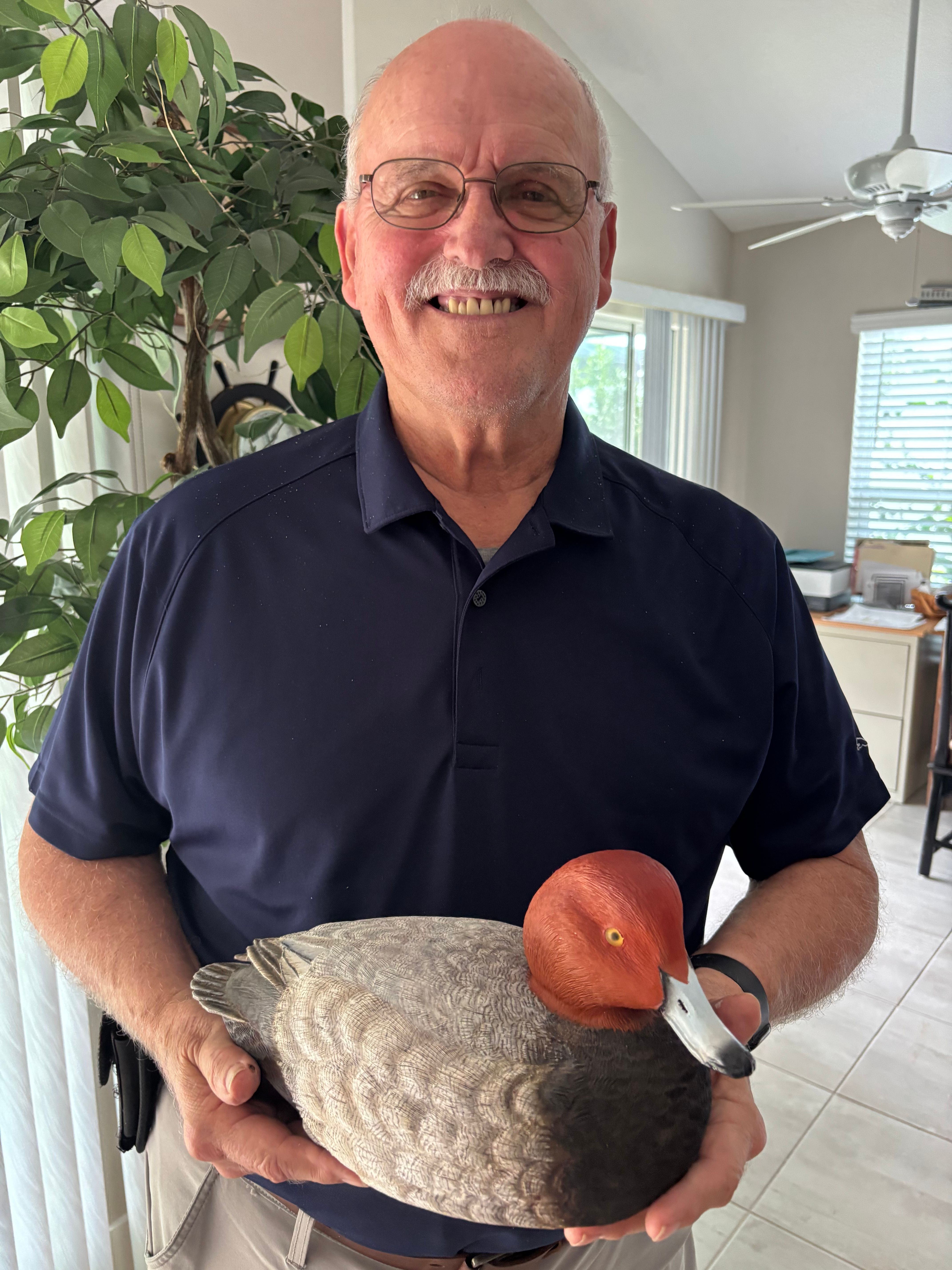 Bill Dodd who has a grey mustache and gold rimmed glasses, clothed in a dark short sleeved shirt is holding a duck carving in his two hands.