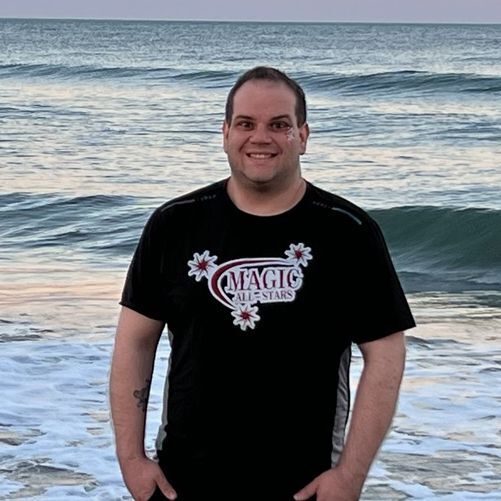 A young man in a black short sleeved tee shirt standing outside with the ocean behind him