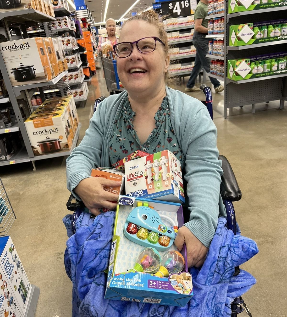 A woman sits in a wheelchair in a store aisle, looking slightly off camera, smiling, holding toys in her lap