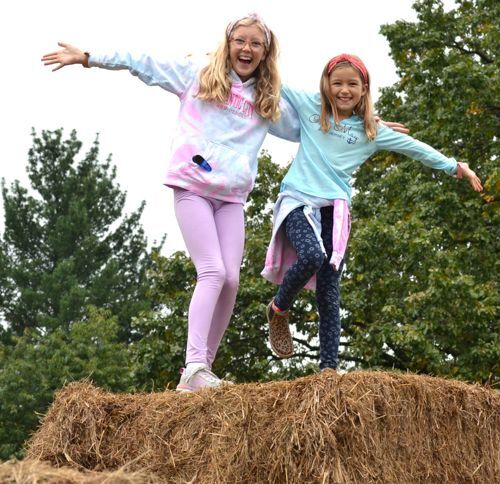 2 girls hold their arms out wide and shout with joy at the top of a straw pyramid