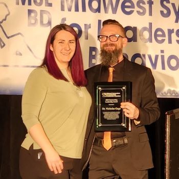 A smiling woman with shoulder-length red hair and a light green top stands beside a bearded man in a black suit and orange tie holding a black-framed award plaque. They pose in front of a “Midwest Symposium for Leadership in Behavior Disorders” banner.