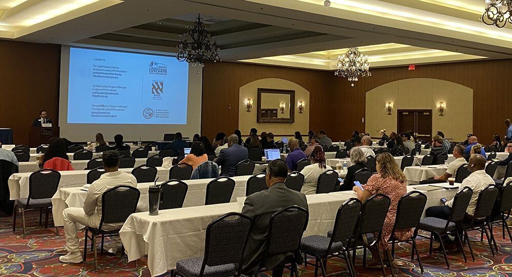 Conference room with attendees seated at tables, facing a speaker and projected presentation slide.