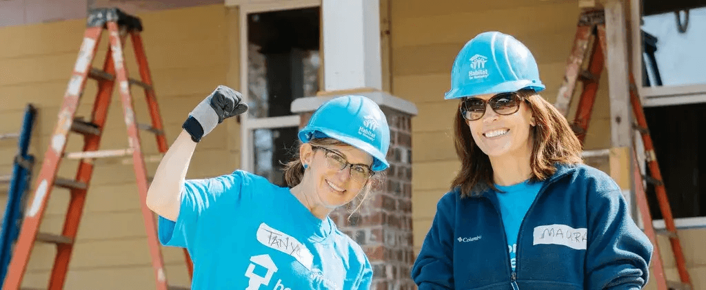 Two smiling female Habitat for Humanity volunteers in blue hard hats and shirts pose in front of a home under construction. One woman raises her fist in a cheerful, empowered gesture.