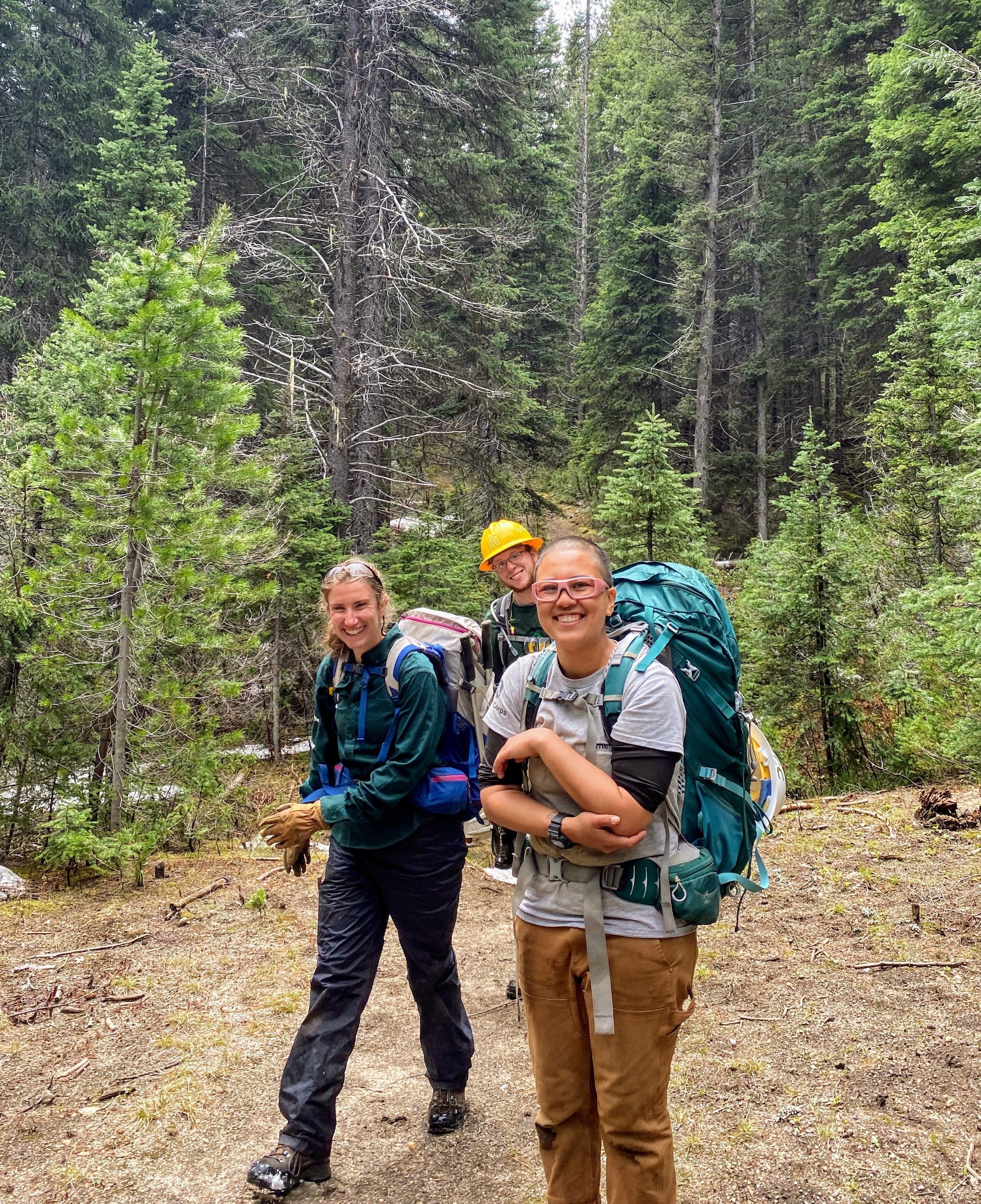 Three people are smiling towards the camera in a dirt clearing. Trees line the clearing in the background.