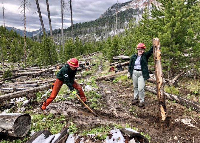 [Image Description: Two MCC members are installing a trail structure called a water bar. One member is digging the trench for the log, while the other holds the log and poses for the photo.]