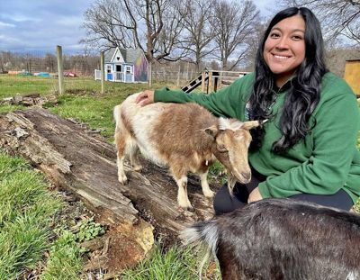 A woman sits on the end of a log petting a baby goat standing on the log to her right.