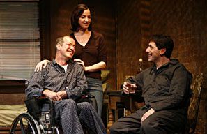 THE RULES OF CHARITY - 2007. Pamela is standing behind Christopher and she is placing her hands on his shoulders. Christopher is sitting in a wheelchair looking at Nick. Nick is sitting on a chair and looking at both of them.