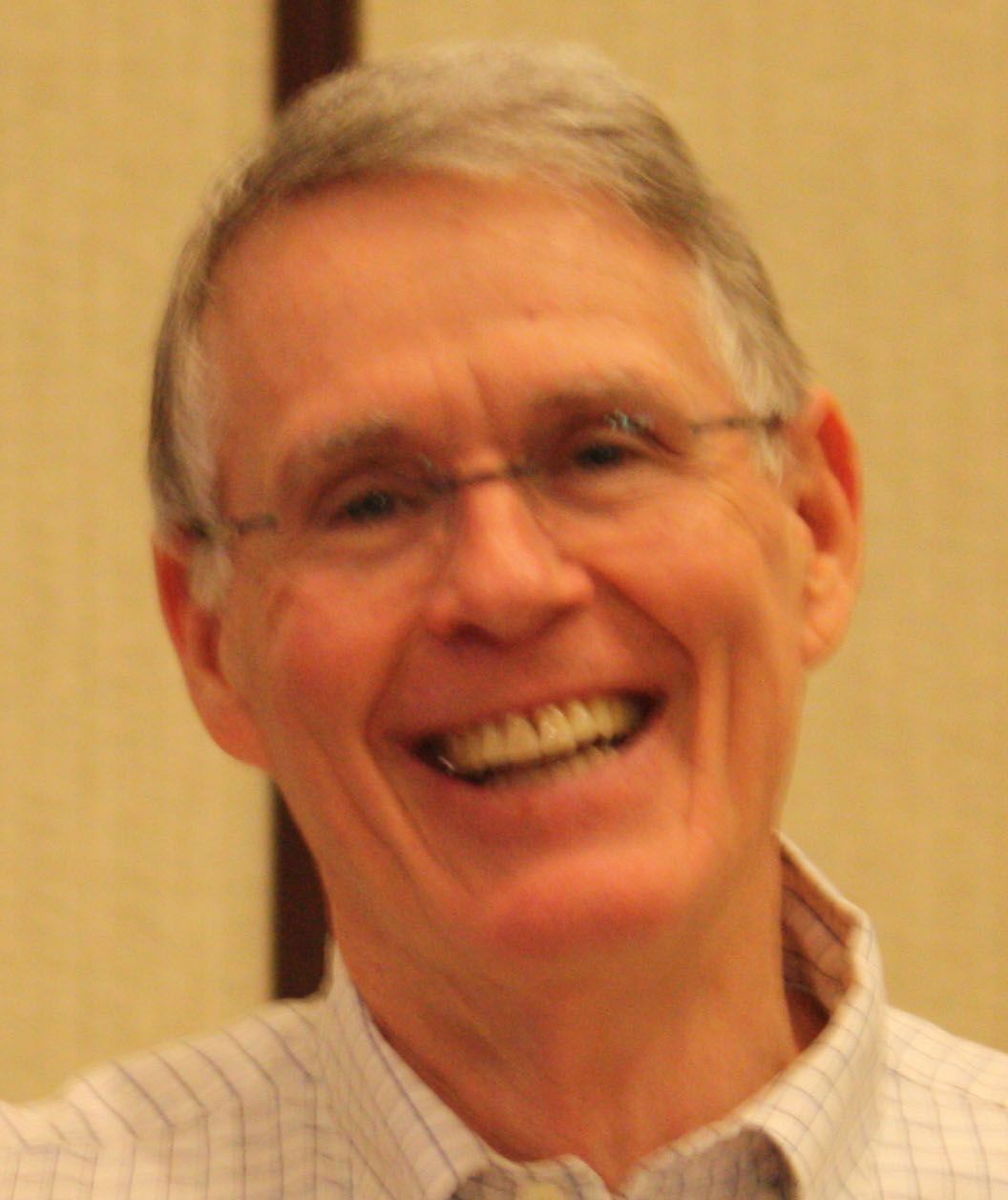 Close-up of an older man with short gray hair and rimless glasses smiling broadly. He wears a light checkered button-up shirt and stands against a neutral indoor backdrop. His warm expression and visible teeth convey genuine happiness.