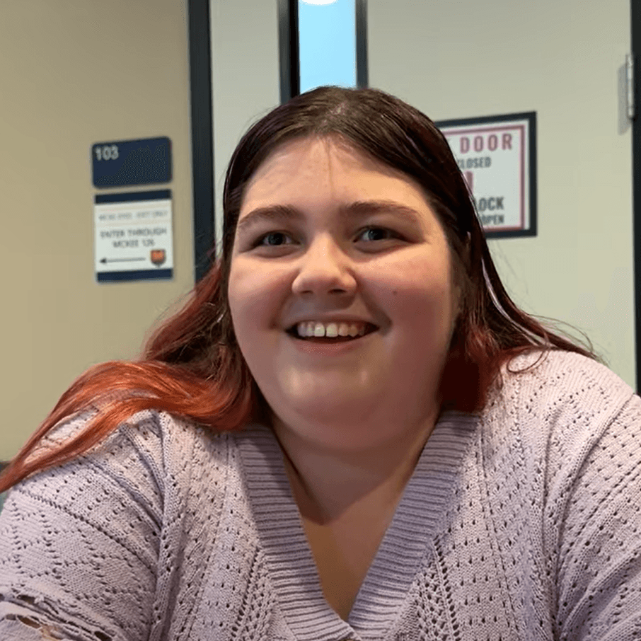 Desiree is a white woman with brown hair that is died red at the bottom. In this photo, she wears a light purple sweater and smiles for the camera.