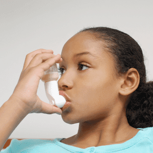 Photo of a young girl using an inhaler for asthma. The inhaler is made of white plastic and is held in her hand, which presses it against her lips. The girl is facing the left and wears a light teal shirt.