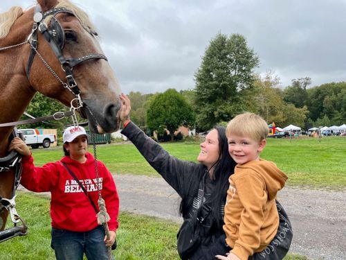 A mother holds her young son in her left arm while with her right arm she reaches up to pet the nose of a very tall horse.