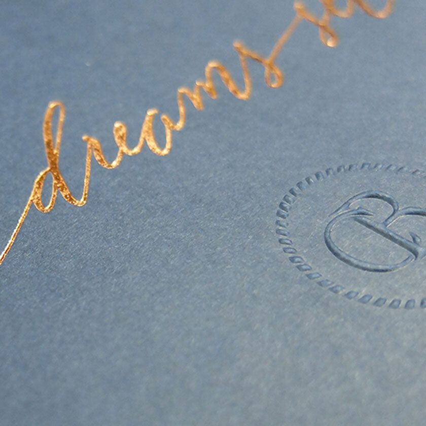 Close-up of blue paper with gold foil cursive lettering and an embossed circular logo.