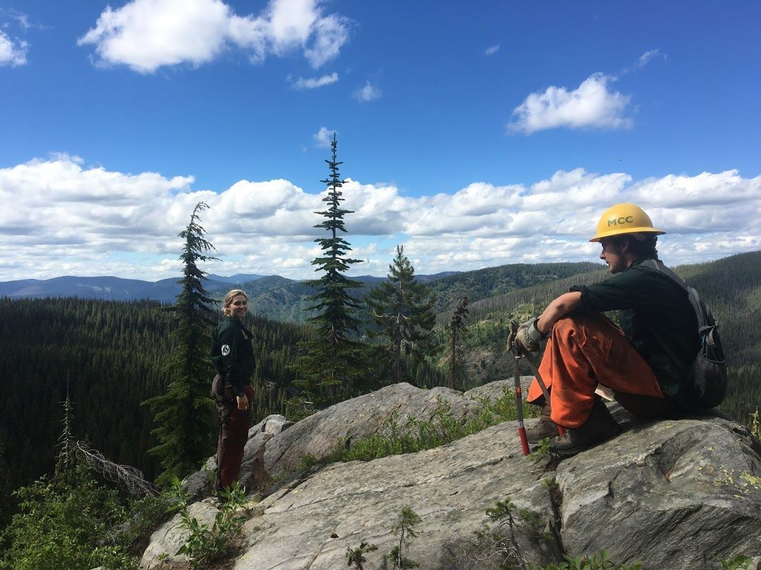 [Image Description: Two MCC members taking a brief break; one is sitting on a rock, the other is standing nearby. They are both in their uniforms, looking out at the expansive, mountain view surrounding them.]