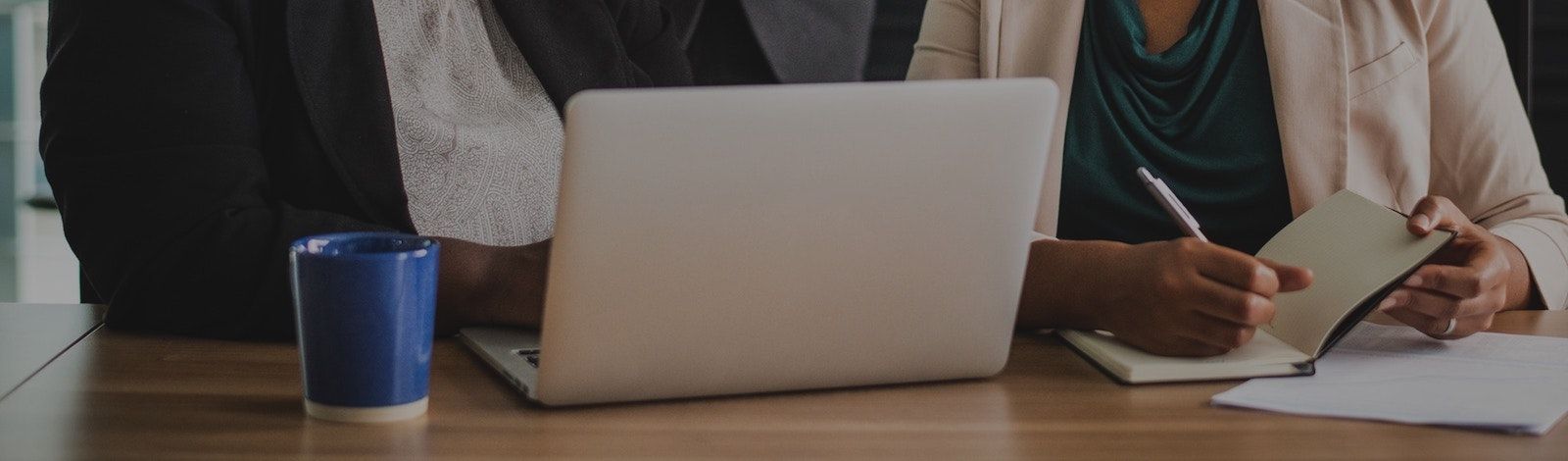 Foto de dos mujeres de negocios sentadas hombro a hombro en una mesa, detrás de un laptop plateado. La mujer a la izquierda lleva una camisa blanca bajo una chaqueta negra. La mujer a la derecha lleva una camisa verde azulado bajo una chaqueta crema y est