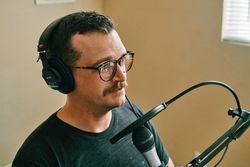 A seated man with moustache talking into a microphone