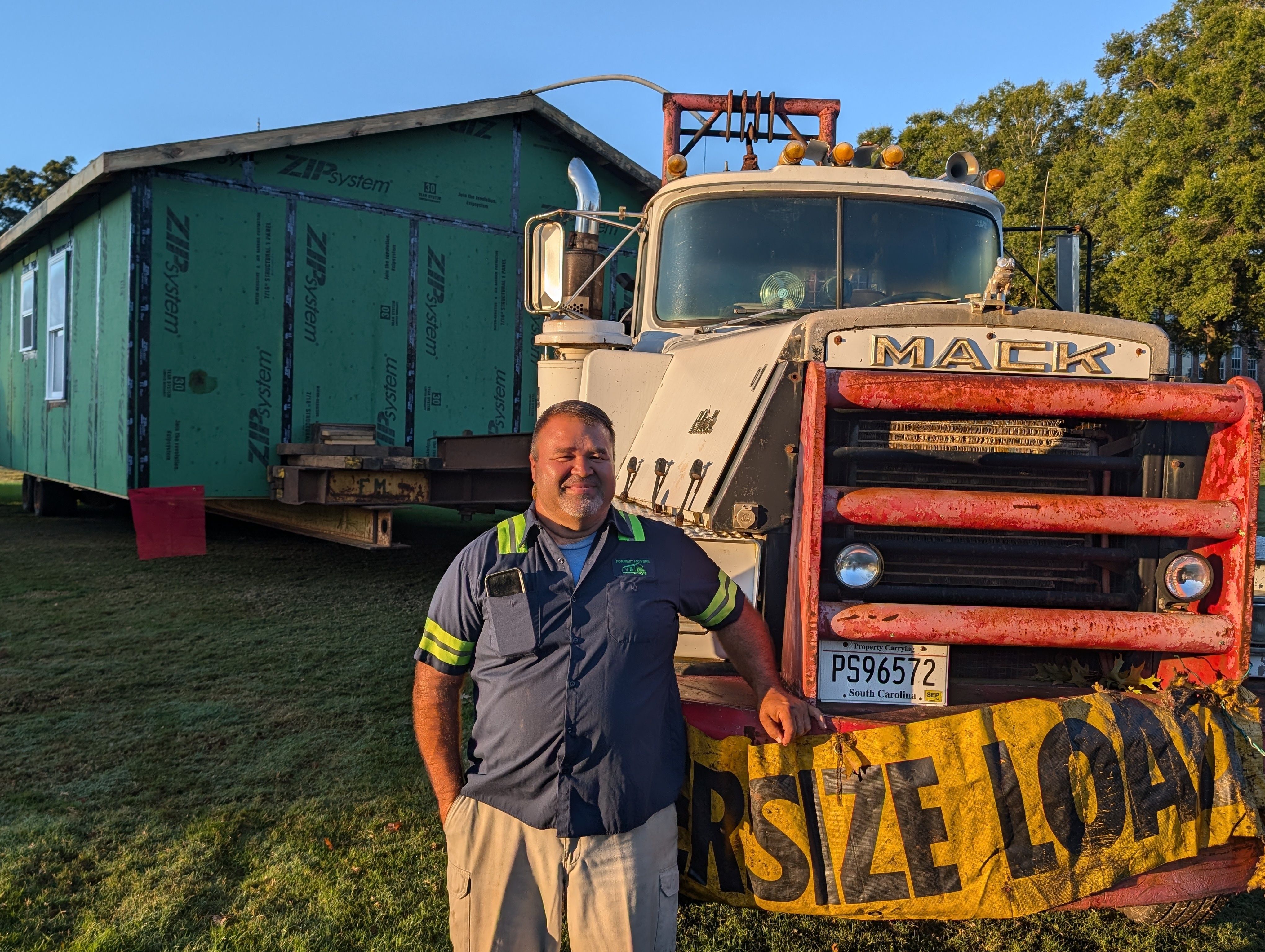 George Forrest of Forrest Movers shown standing next to a truck with an overside load sign and the Homecoming House loaded on the back