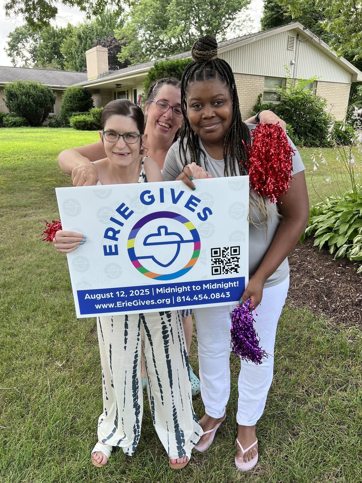Three smiling women stand outside holding a sign promoting Erie Gives Day