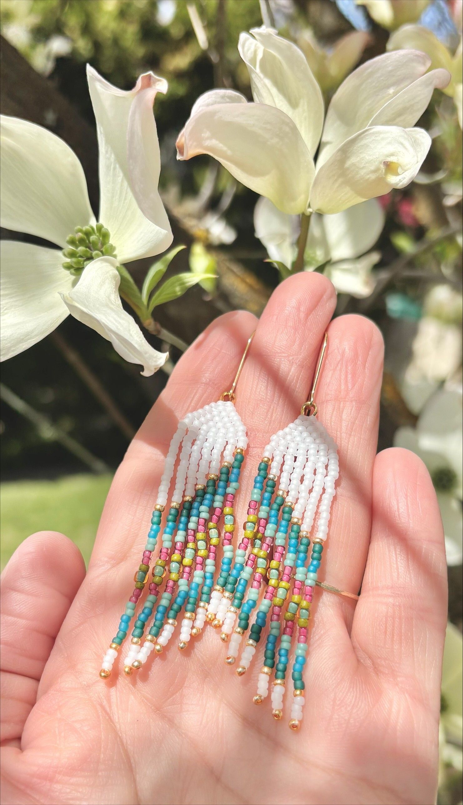A pair of fringed earrings with stripes of white, green, blue, pink, and gold sits in the artist's hand with white flowers behind. 