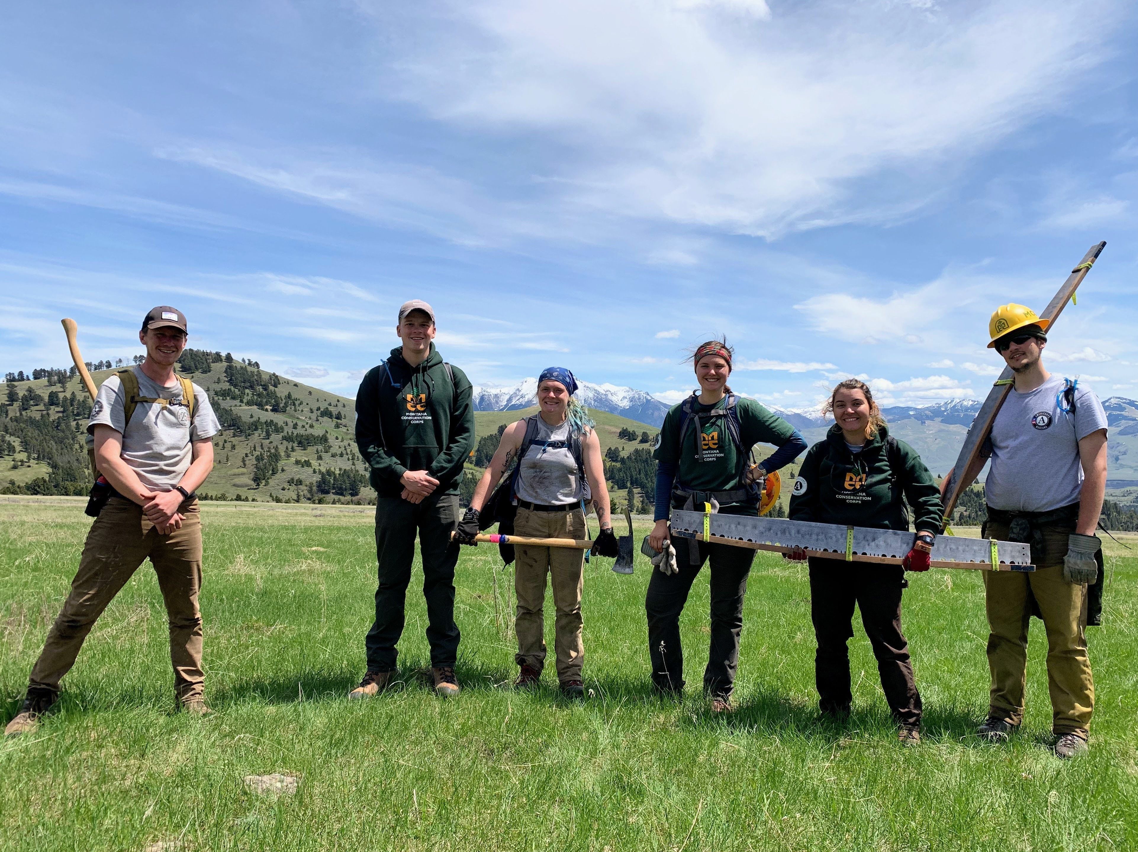 [Image description: Six MCC members stand in a field, two holding large crosscut saws, and two with mattocks in their hands.]