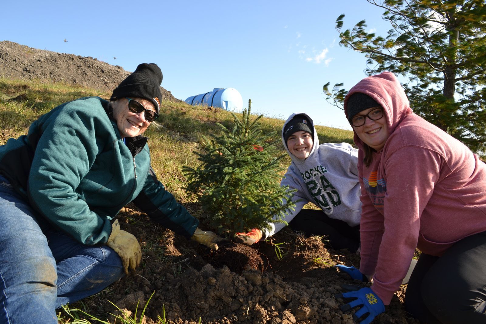 Three women pose next to an evergreen tree that has been recently planted. 