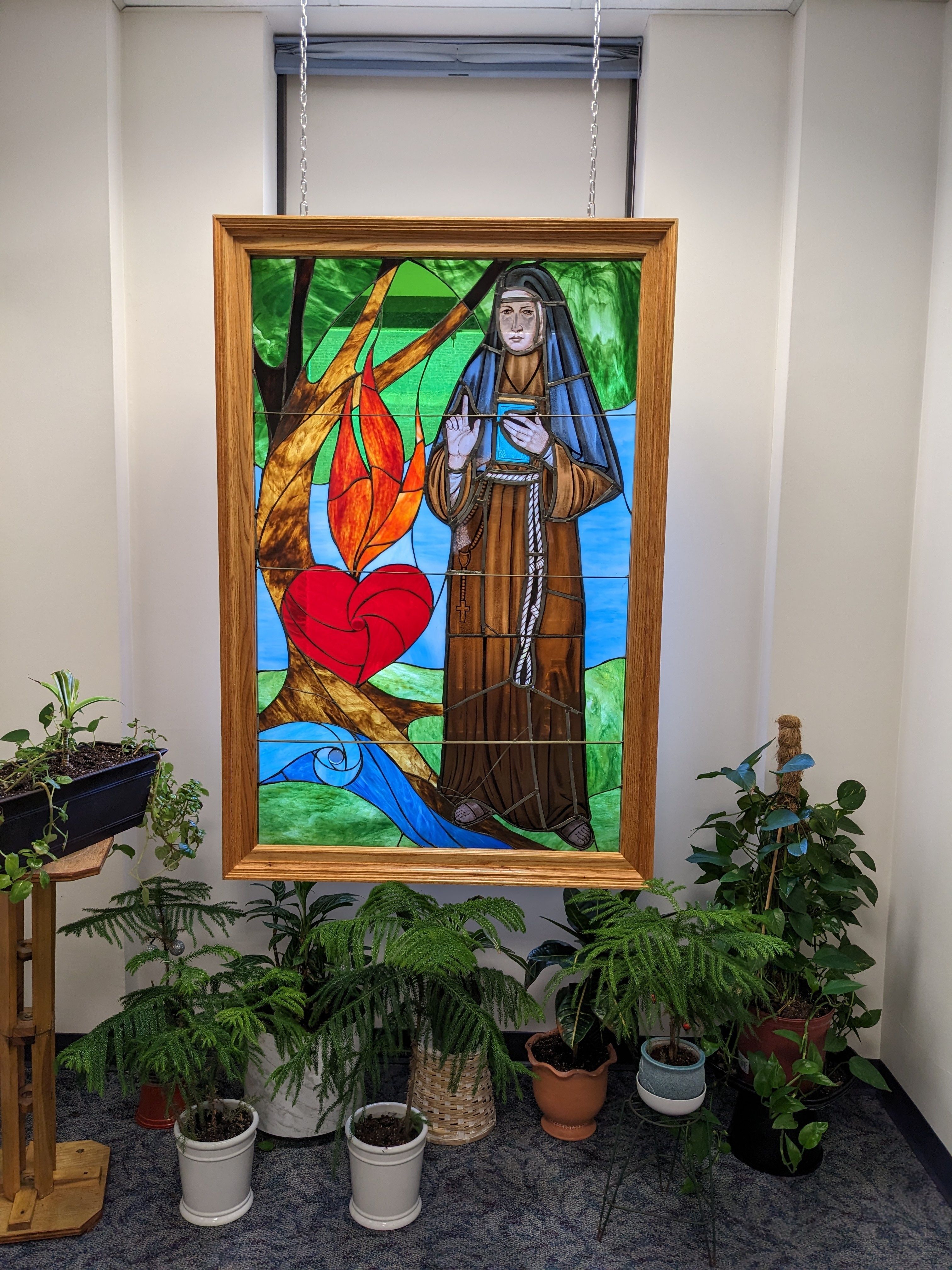 A framed stain glass window of Blessed Mary Angela, founder of the Felician Sisters, hangs on the wall above green potted plants. 