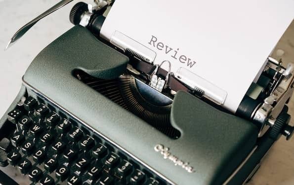 A side-angled photo of an old fashioned grey typewriter. A white piece paper with the word "Review" sticks out of it.