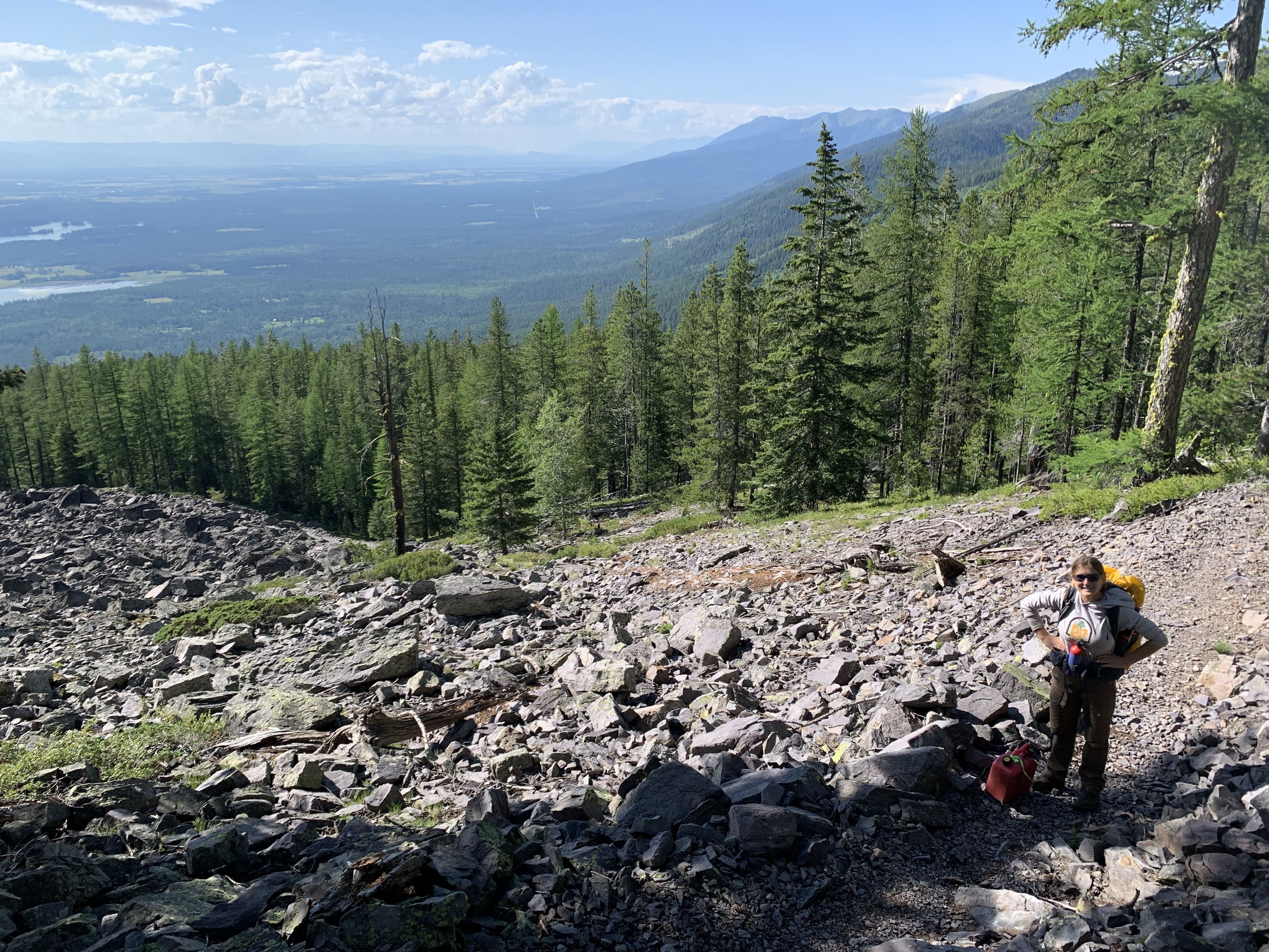 A crew member stands on a rocky scree slope, with a view of a mountain valley and a large lake below.