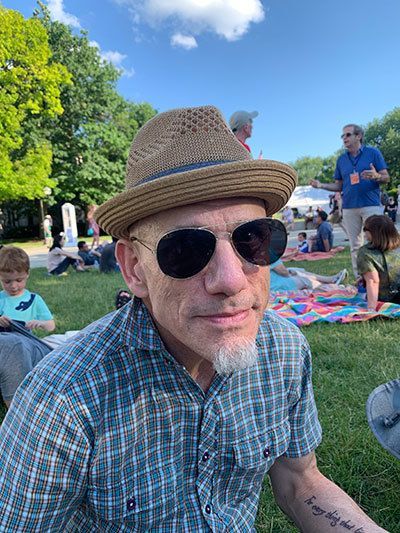 Dr. Ruderman is sitting outside on a nice day wearing aviator sunglasses and a stylish hat.
