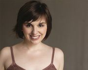Bree Klauser (They/Them/Theirs), a person who is in their 20's-30's. They are wearing a brown blouse, looking straight at the camera smiling with a dark back drop.