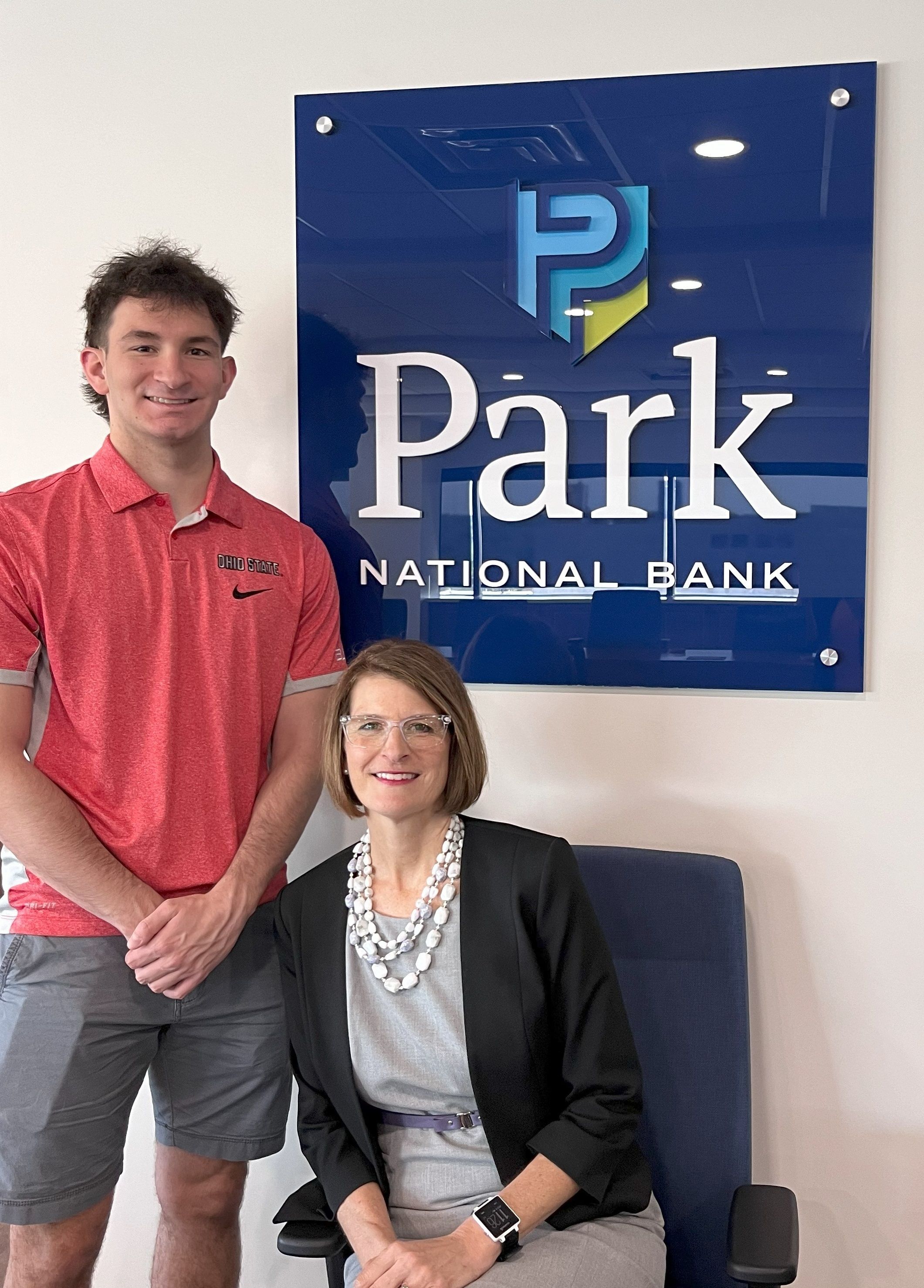 A young man stands beside a seated women in front of a sign that says "Park National Bank"