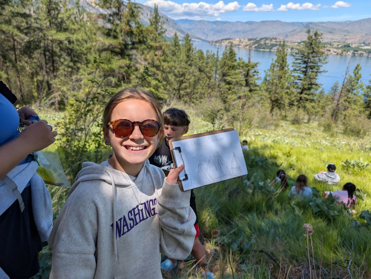 A girl holding up her artwork in front of scenic Lake Chelan during a Field Day.