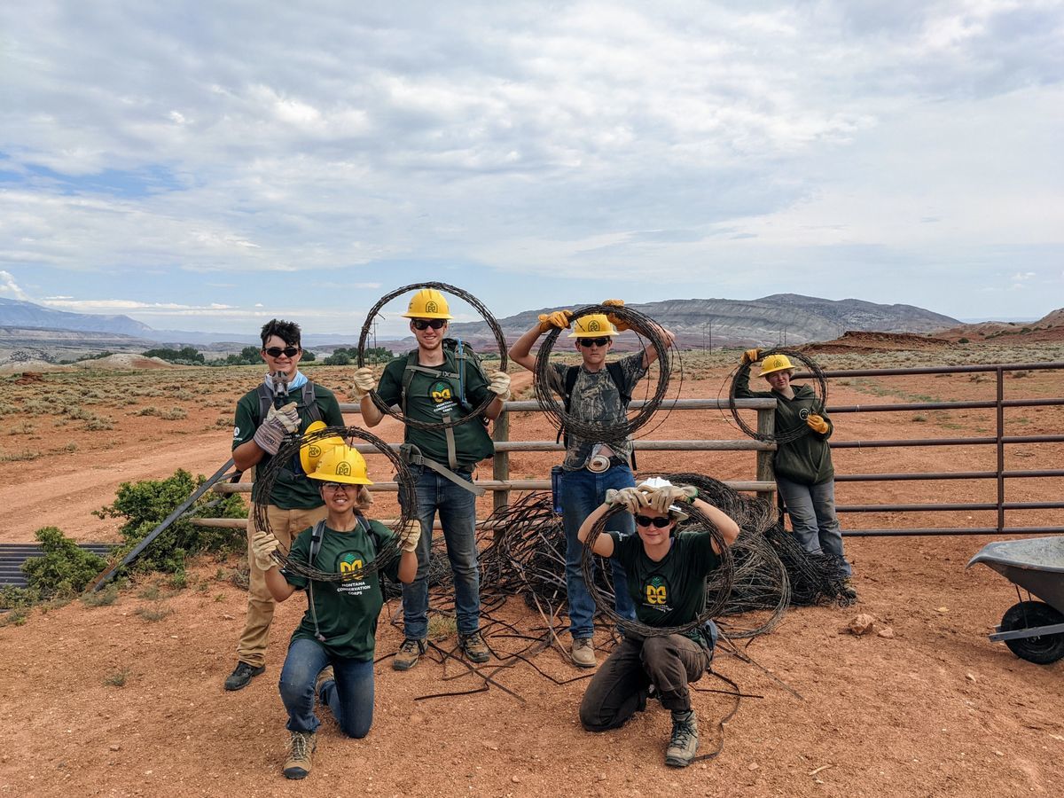 [Image Description: Six MCC Youth members holding barbed wire loops that they used to install fencing. All members are wearing hard hats, wearing large welding gloves, and smiling.]