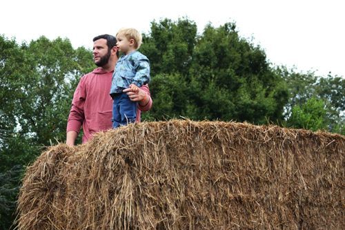 A man holds his young son up on top of a bale of straw. This bale of straw is at the top of the straw pyramid and they are both looking off to the left at something far in the distance.