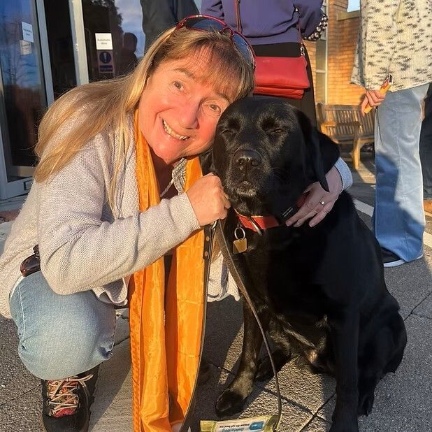 A smiling Issy - long blond hair with bangs, sunglasses on her head, wearing blue jeans, a sweater and scarf - kneels down next to her black lab guide dog.
