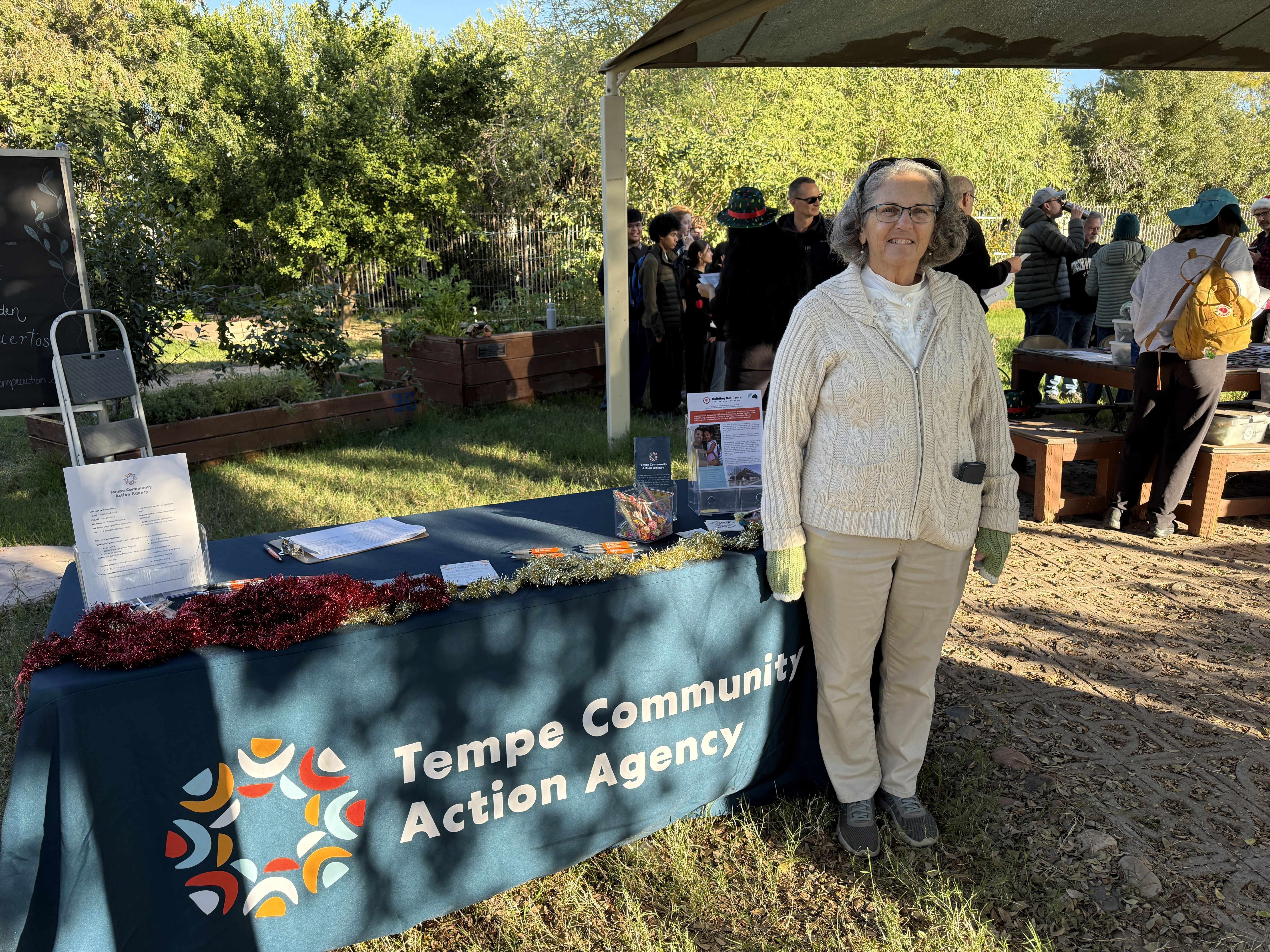A woman in beige standing next to table in a garden. In the background are volunteers gathering.
