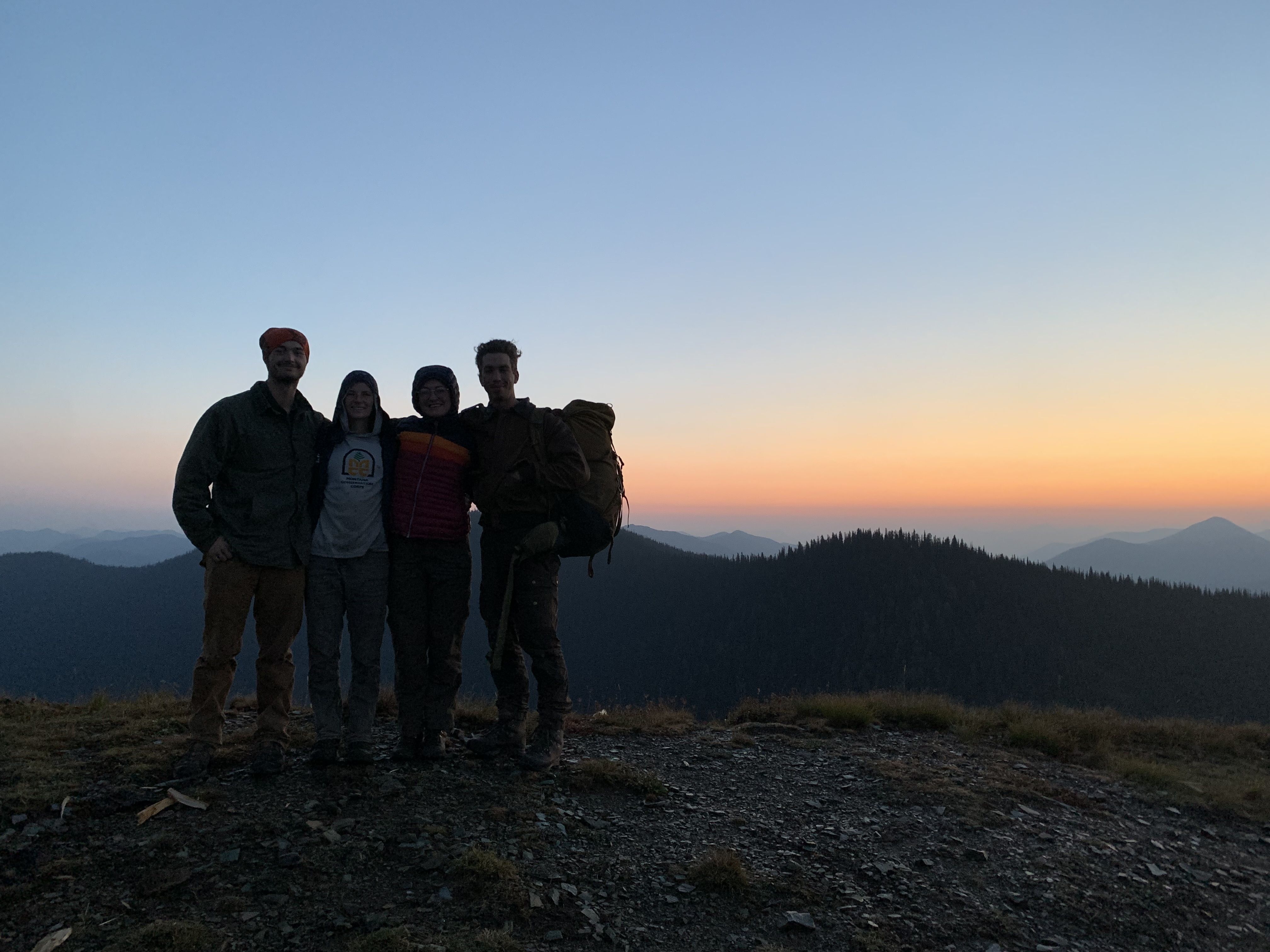 A crew stands on a hill top, with the sun setting behind them.