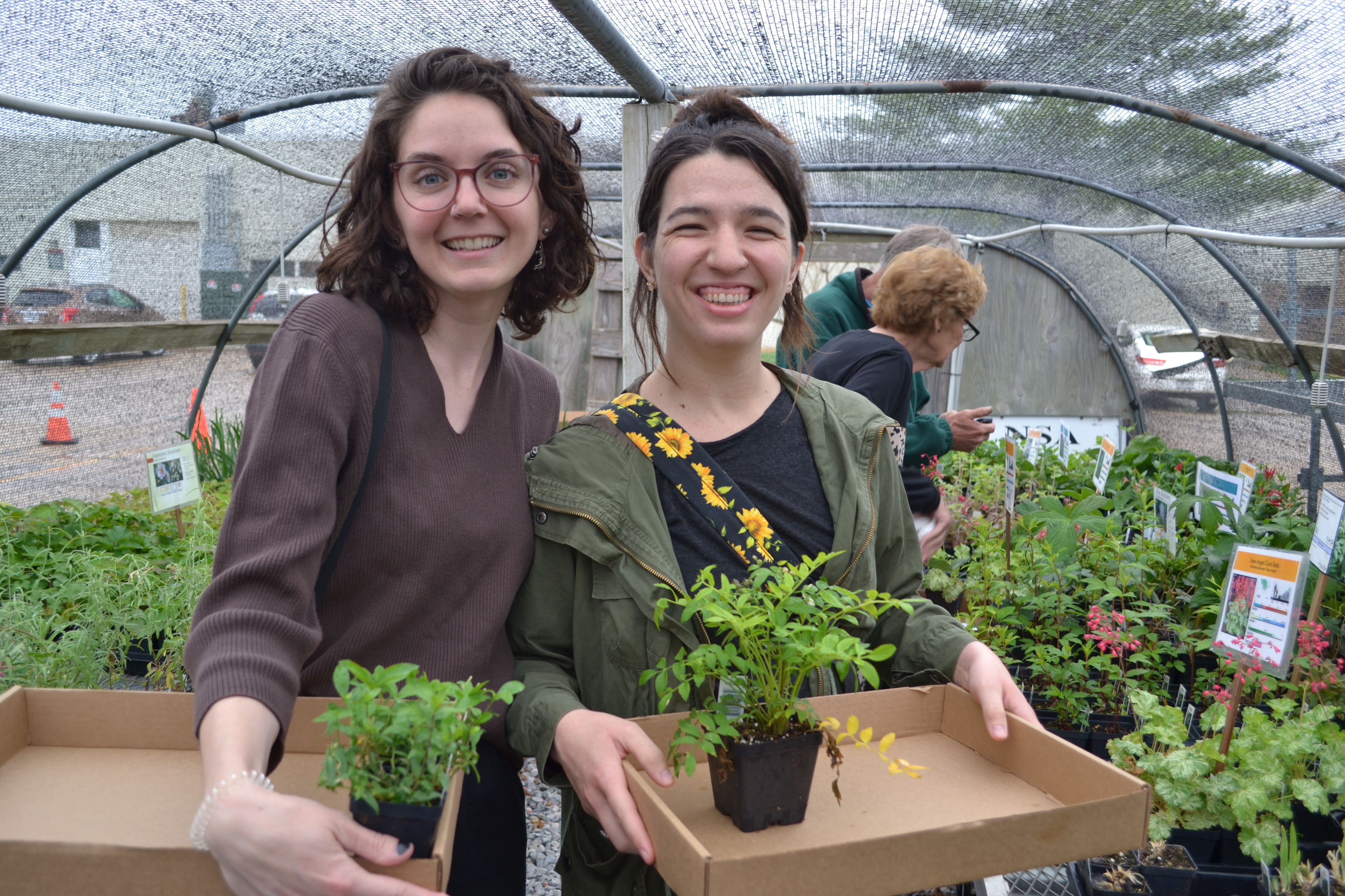 A young woman wearing a brown shirt stands next to another young woman wearing a black shirt and cardigan, holding a flat of plants in a greenhouse 