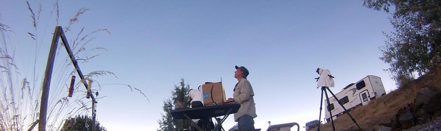 Person setting up equipment outdoors near a telescope and RV at dusk.