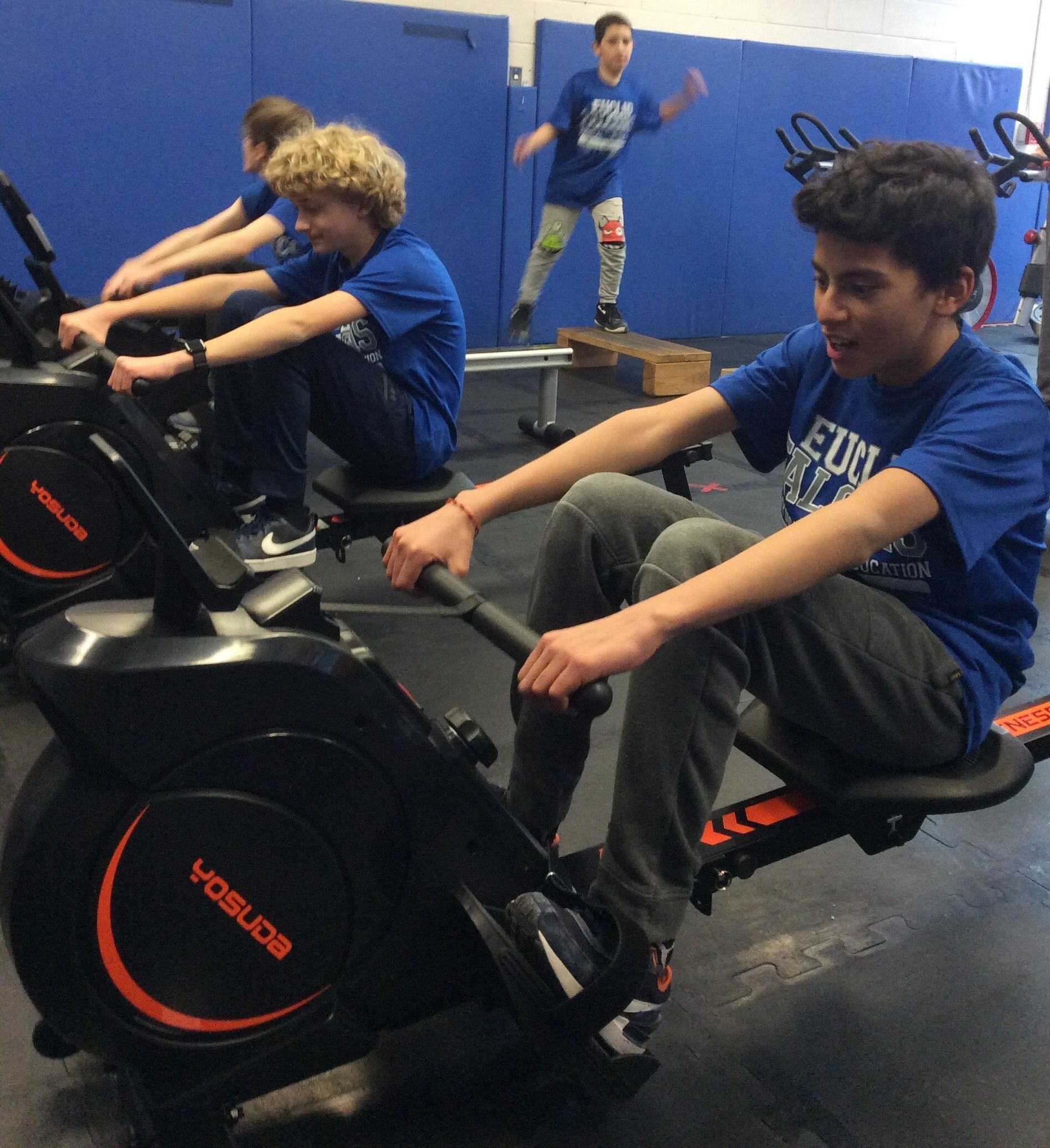 Two middle school students working out on a rowing machine. One has blonde hair. One has dark hair.